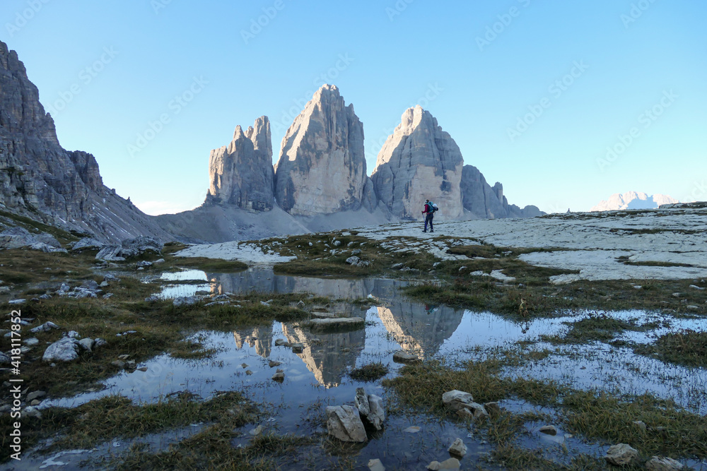 Obraz premium A woman enjoying the view on the Tre Cime di Lavaredo (Drei Zinnen), mountains in Italian Dolomites. The mountains are reflecting in small paddle. Desolated and raw landscape. Early morning. Daybreak