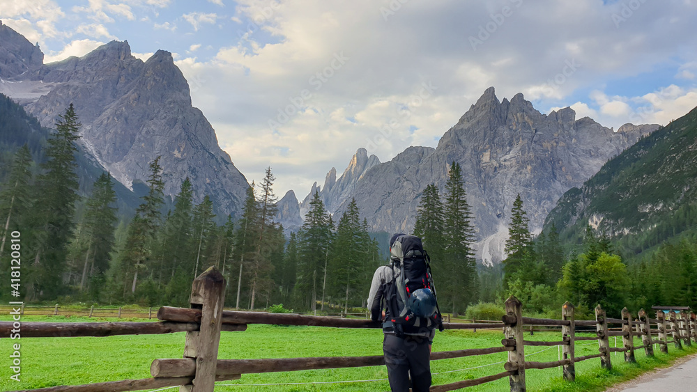 A man with a big hiking backpack enjoying the distant view on Italian Dolomites. There is a lush green meadow with a wooden fence around it. Morning fog in the valley. Thick forest on the slopes