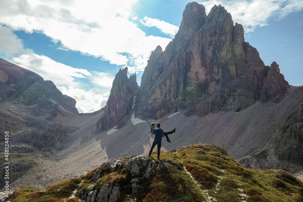 A man holding a woman in his arms under the foot of high Italian Dolomites and enjoying the panoramic view. There are many sharp peaks in front. Lots of lose stones and landslides. Sharing experience.