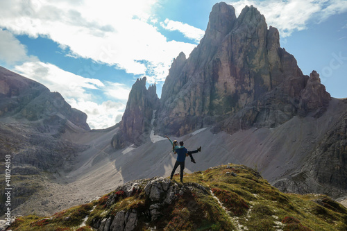 A man holding a woman in his arms under the foot of high Italian Dolomites and enjoying the panoramic view. There are many sharp peaks in front. Lots of lose stones and landslides. Sharing experience. © Chris