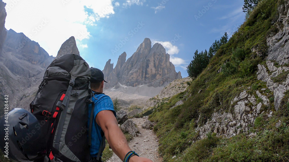 A man with big backpack taking a selfie while hiking along a narrow pathway in Italian Dolomites. There is a massive mountain with very steep and sharp slopes. Smaller mountains around. Raw landscape