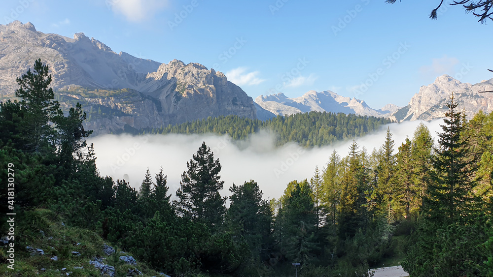 An early morning in Italian Dolomites. A thick forest overgrowing the area. The valley is shrouded in morning haze. There are high, stony mountain chains around. Golden hour. New day beginning