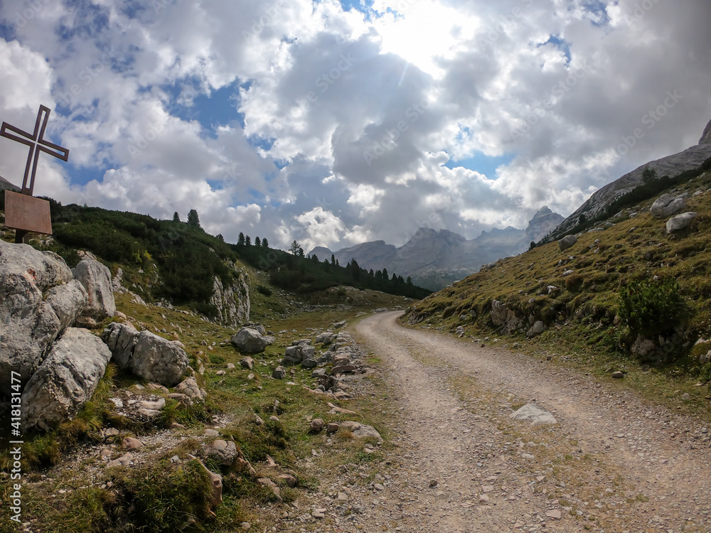 A gravelled road leading through Italian Dolomites. There is a metal cross on the boulder next to the road. High mountains around. The slopes are green. Overcast. Spirituality and calmness.