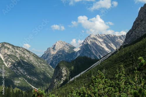 A lush green valley in Italian Dolomites. There are tall trees and high mountains around. The mountain slopes are rocky, with small plants overgrowing them. Dense forest on the lower slopes. Sunny day © Chris