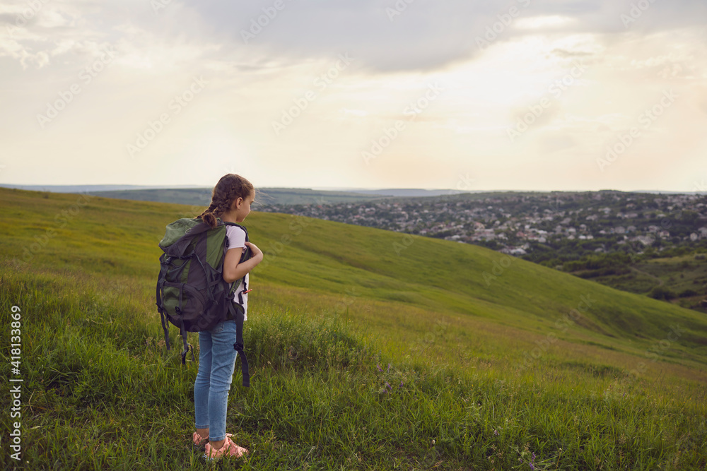 Girl tourist hiker with backpacks looks at the sunset standing on a hill in the summer. Little kid girl on foot travel adventure in nature