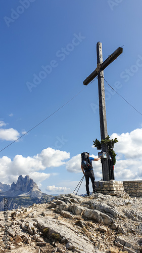 A man with a big hiking backpack standing under a massive wooden cross at the top of  Strudelkopf in Italian Dolomites. Drei Zinnen in the back. The man enjoys the view. High Alpine landscape. Freedom photo