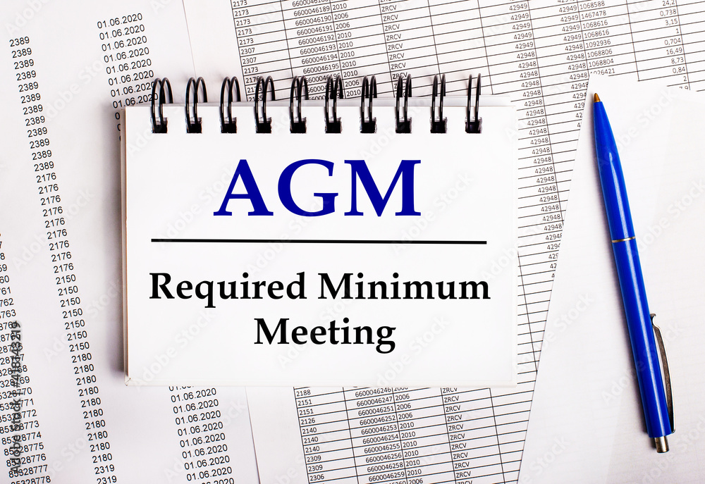 On the table are charts and reports, on which lie a blue pen and a notebook with the word AGM Annual General Meeting On the table are charts and reports, on which lie a blue pen and a notebook with the word AGM Annual General Meeting