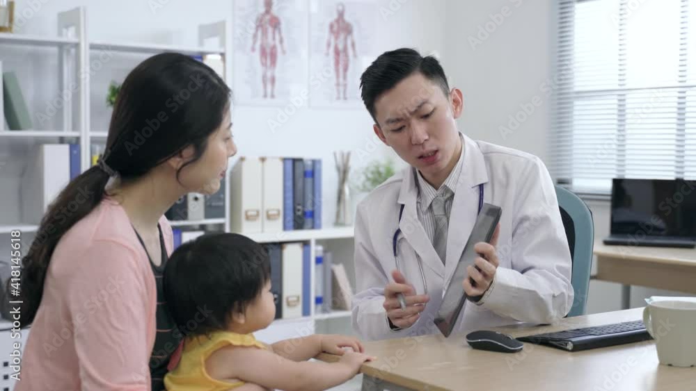 asian pediatrician is tapping the screen with a pen while explaining the test results to the mother and her kid on a touchpad in his clinic office. asian pediatrician is tapping the screen with a pen while explaining the test results to the mother and her kid on a touchpad in his clinic office.