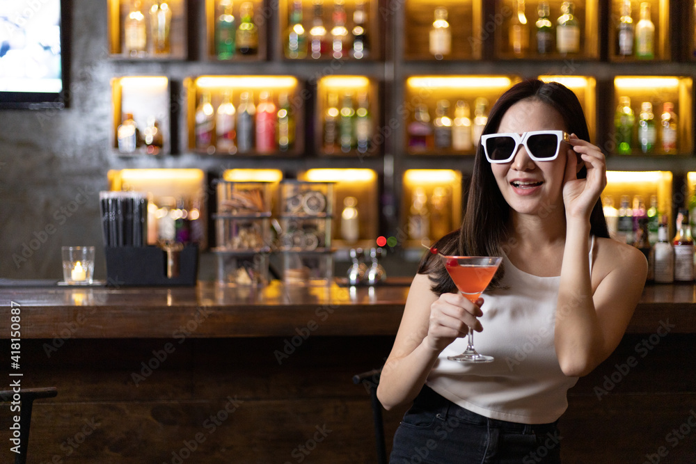 Young Asian woman with black glasses enjoys drinking a cocktail in front of a vintage bar, Relaxing activities after work or hangouts, Place of entertainment for young adolescents or night club party.