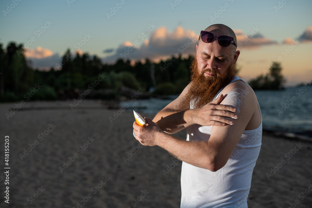Bearded man applying sunscreen on body while standing on the beach. Bearded man applying sunscreen on body while standing on the beach.