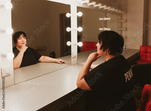 A plump brunette sits in front of a mirror with light bulbs in a dressing room in the dark