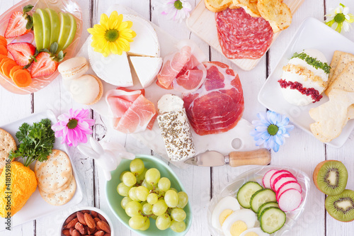 Spring or Easter theme charcuterie table scene against a white wood background. Assortment of cheese, meat, fruit and vegetable appetizers. Above view.