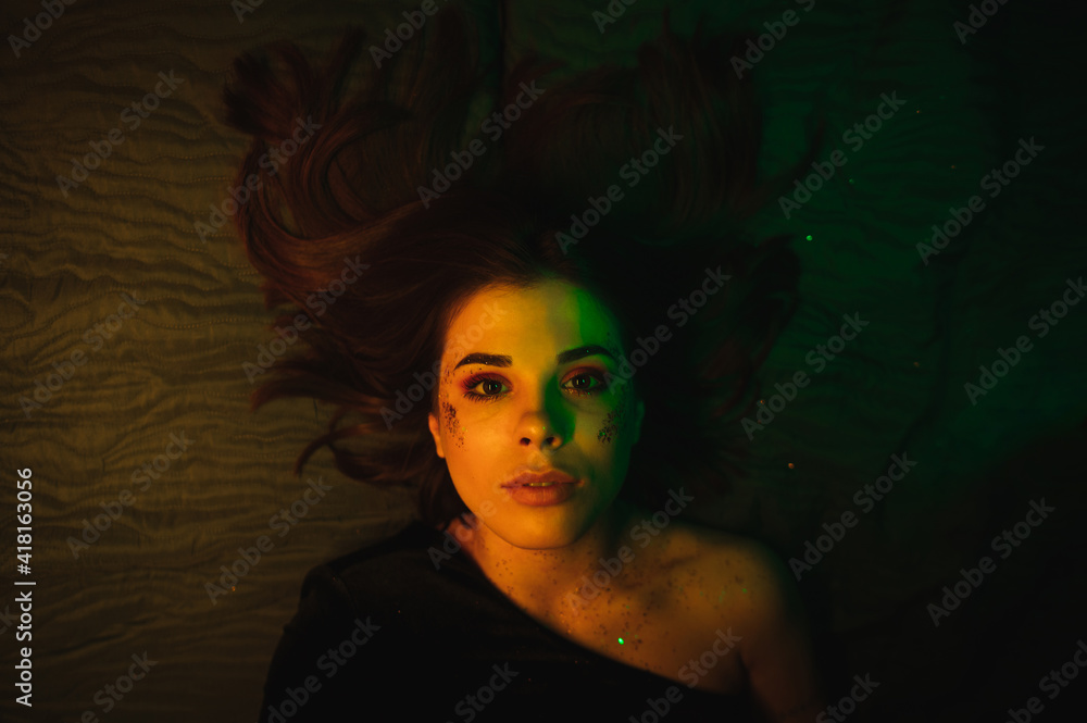 Beautiful woman with bright makeup with glitter on her face and in a black dress lies on the bed after the party and looks at the camera with a serious face in the orange and green light. Top view
