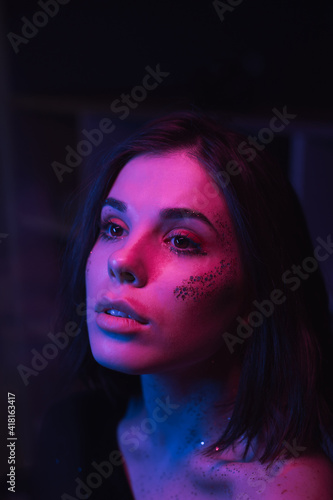 Stylish cinematic portrait of a brunette woman with glitter on her face and beautiful makeup in a room with purple and blue light, looking away.