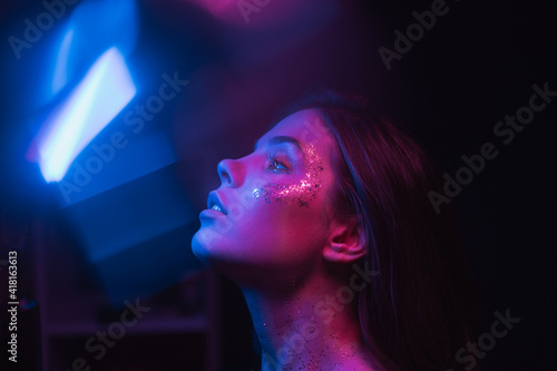 Blurred night art portrait of a woman in neon. Portrait of a skinny brunette with glitter on her face in purple and blue light, looking to the side and glare