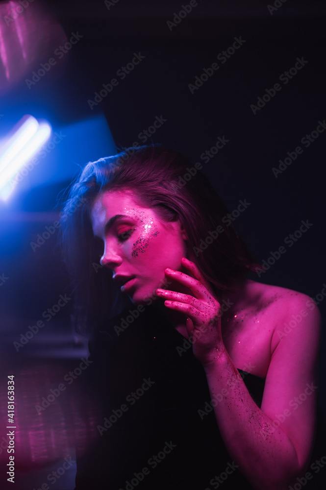 Neon fashionable night portrait of a woman with glitter on her face and bright makeup at a party with colored light. Vertical