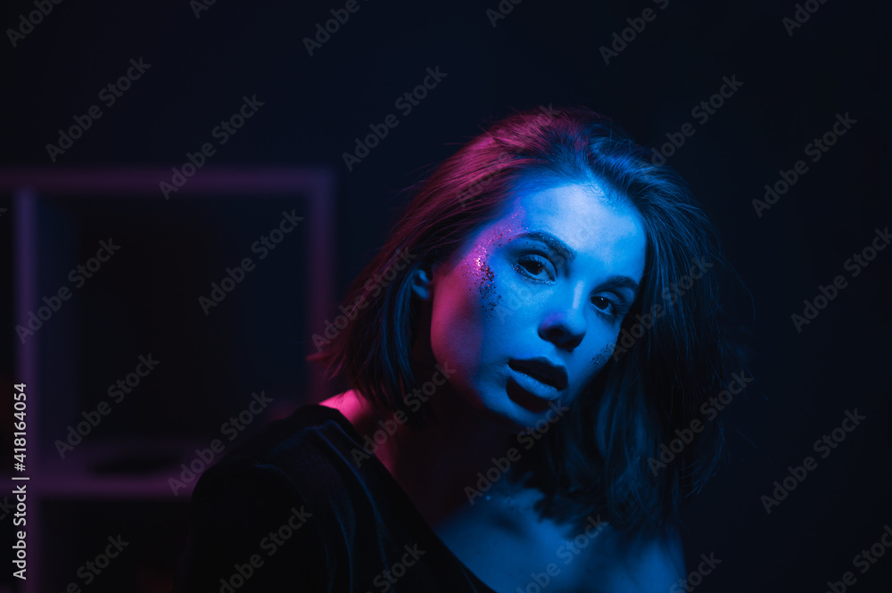 Night neon portrait of a stylish beautiful woman with makeup with glitter in blue and purple light on the party, looking at the camera with a serious face.