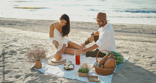 Beautiful couple having romantic breakfast with lots of tasty food and wine, sitting on piknic planket at beach with ocean view photo