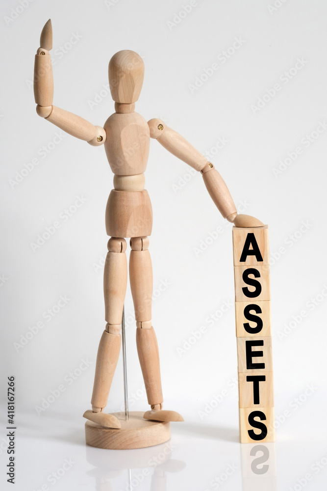 The wooden man raised his hand, and under his left hand he has cubes with the inscription - ASSETS The wooden man raised his hand, and under his left hand he has cubes with the inscription - ASSETS