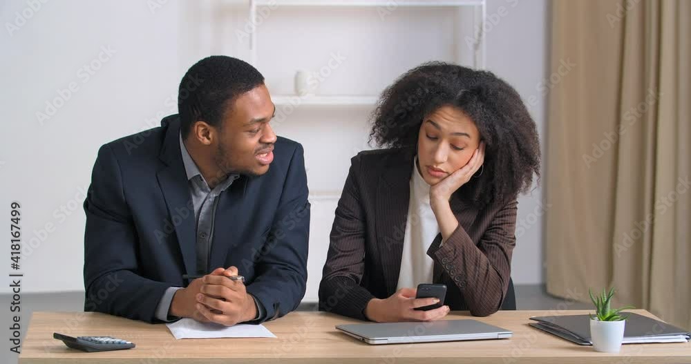 Two colleagues sitting at business meeting ethnic woman manager bored looks at phone chatting online on social networks afro american man snaps his fingers needs attention calling female coworker Two colleagues sitting at business meeting ethnic woman manager bored looks at phone chatting online on social networks afro american man snaps his fingers needs attention calling female coworker