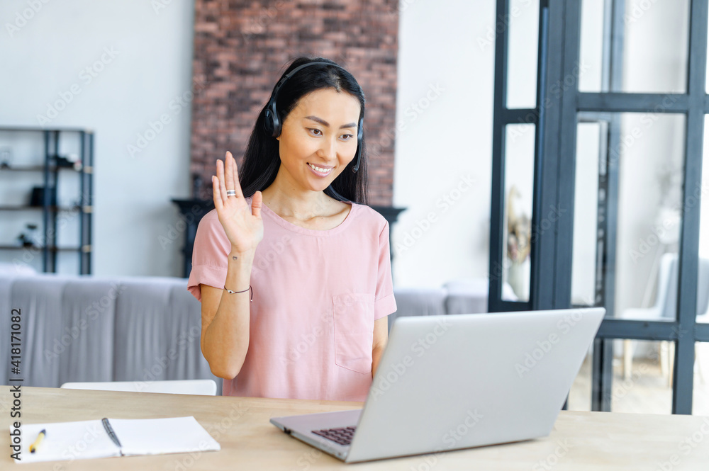 Brunette Asian woman wearing headset using a laptop for video communication, smiling female student waving hi into a webcam, has online video meeting, participates in the webinar Brunette Asian woman wearing headset using a laptop for video communication, smiling female student waving hi into a webcam, has online video meeting, participates in the webinar