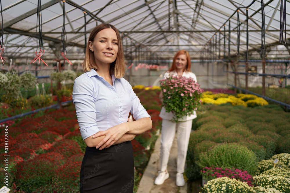 Smiling Greenhouse owner posing with folded arms having many flowers in background and a colleague holding a pot with pink chrysanthemums under glass roof