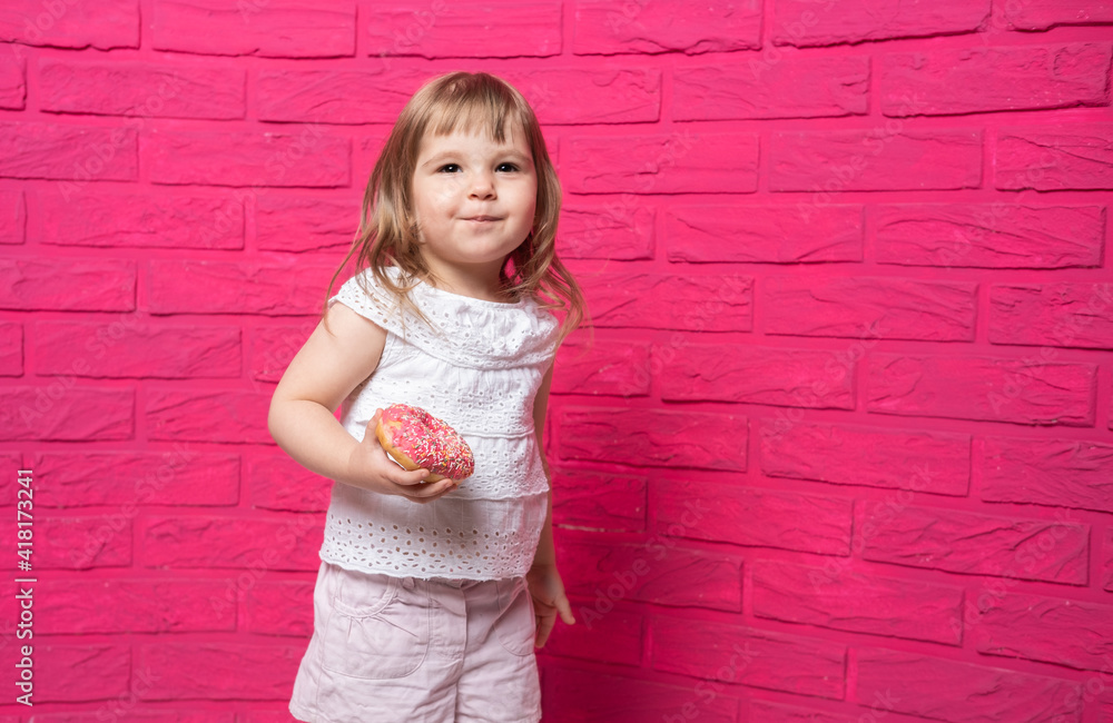 little funny blonde girl in white shirt eats donut on pink background. little funny blonde girl in white shirt eats donut on pink background.