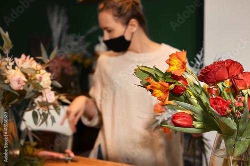 Florist works with colors. Flower seller chooses flowers for future bouquet. Flowers shop worker in a mask standing in flower shop and checking flowers in glass vase.