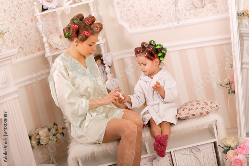 Adorable little girl with her mother in curlers paint their fingernails. Copies mom's behavior. Mom teaches her daughter to take care of herself. Beauty day