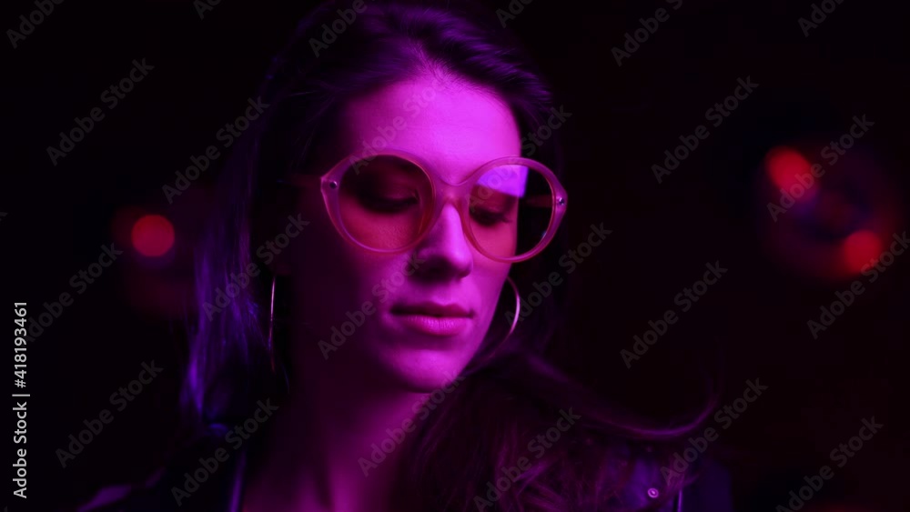 Portrait beautiful trendy girl In glasses looking at the Camera. Attractive Woman in Purple Blue Light Posing at Party. Female look around. Close up in Colourful neon background light. Portrait beautiful trendy girl In glasses looking at the Camera. Attractive Woman in Purple Blue Light Posing at Party. Female look around. Close up in Colourful neon background light.
