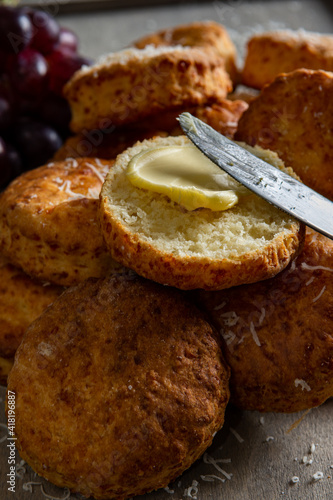 A close up view of a buttered cheese scone and knife positioned atop others and dusted with parmesan cheese. Placed on a grey background with red grapes in the top left of the image.