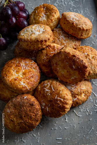 An angled view of a collection of cheese scones dusted with grated parmesan cheese. Placed on a grey background with red grapes to the top left of the image.