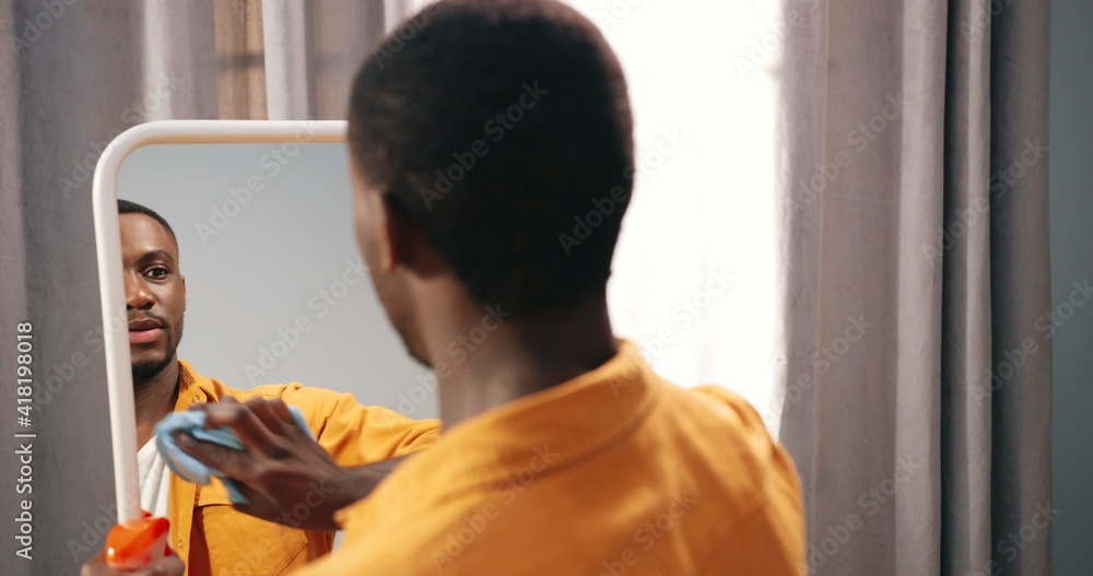 Rear, over shoulder view of handsome happy young African American guy standing in apartment in living room cleaning mirror with glass spray, washing home, disinfecting furniture, housework concept Rear, over shoulder view of handsome happy young African American guy standing in apartment in living room cleaning mirror with glass spray, washing home, disinfecting furniture, housework concept