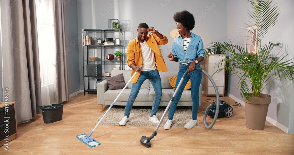 Joyful happy African American young married couple wife and husband cleaning living room and dancing, guy mopping and woman vacuuming floor having fun making funny rhythmic moves in cozy modern home