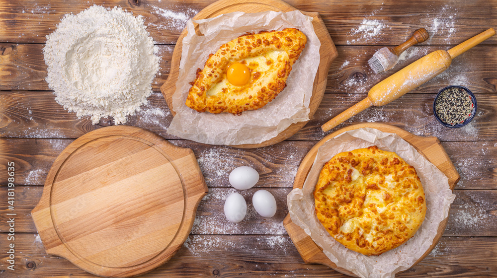Khachapuri lies on wooden round pizza boards, chicken eggs and a rolling pin on a brown board background.