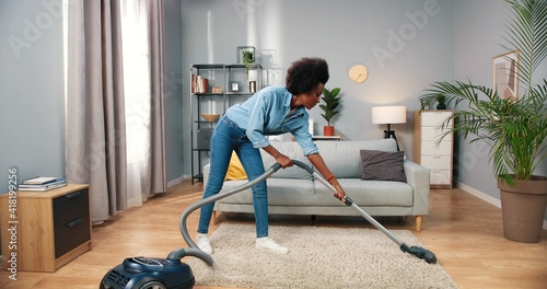 Young beautiful happy joyful African American woman cleaning modern living room vacuuming carpet and dancing having fun playing vacuum cleaner like guitar, housekeeping concept, clean up