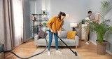 Happy young Caucasian married couple wife and husband clean apartment, woman vacuuming floor in modern room while man wiping dust cleaning furniture in house, family team work, home concept