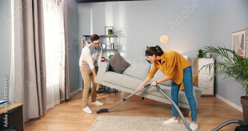 Happy young Caucasian married couple wife and husband clean apartment, woman vacuuming floor in modern room while man wiping dust cleaning furniture in house, family team work, home concept