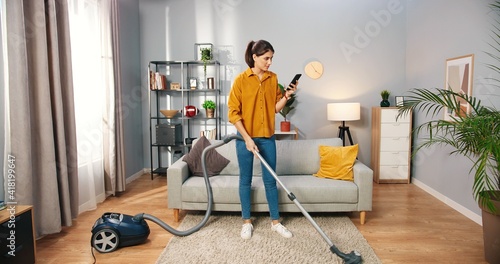 Happy cheerful young Caucasian beautiful woman vacuuming floor and texting surfing internet typing online on smartphone at home in living room, pretty female using vacuum cleaner, domestic concept