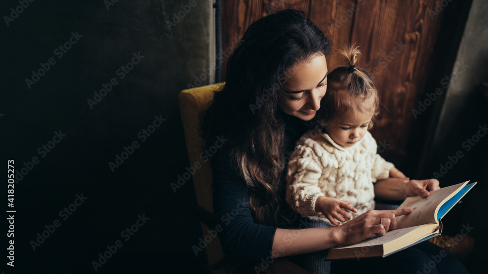 Young mother and her little daughter reading book together sitting in a comfortable chair. Young mother and her little daughter reading book together sitting in a comfortable chair.
