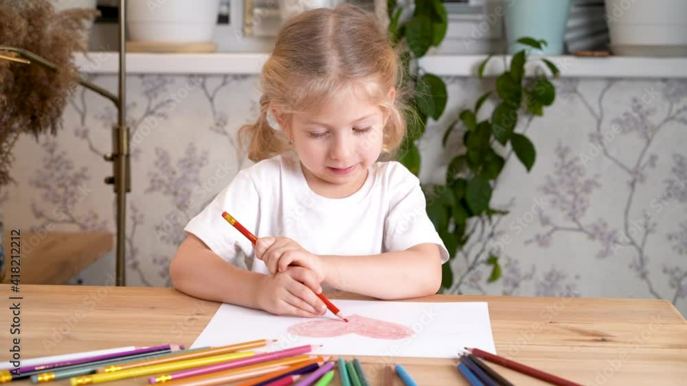 a little girl enthusiastically draws a heart with colored pencils sitting at the table at home alone a little girl enthusiastically draws a heart with colored pencils sitting at the table at home alone