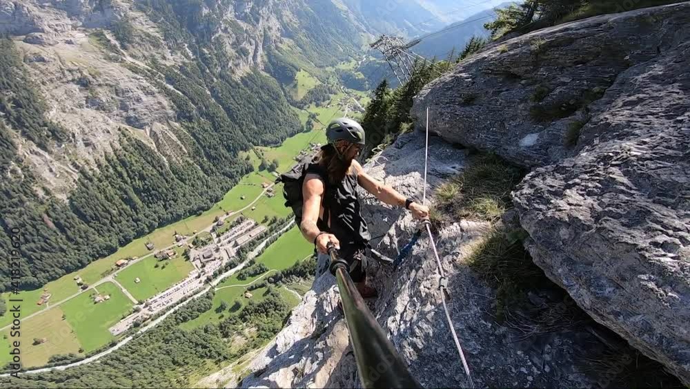 Young man takes a selfie video as he stands on the very narrow and dangerous path on Via Ferrata route in Switzerland with the gorgeous and green valley below during a hot clear summer day. Young man takes a selfie video as he stands on the very narrow and dangerous path on Via Ferrata route in Switzerland with the gorgeous and green valley below during a hot clear summer day.