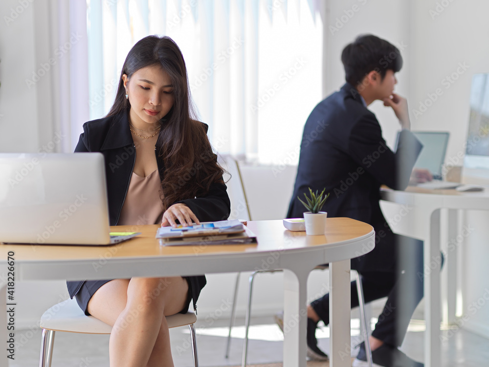 Businesswoman working with laptop and paperwork in office room with coworker in background
