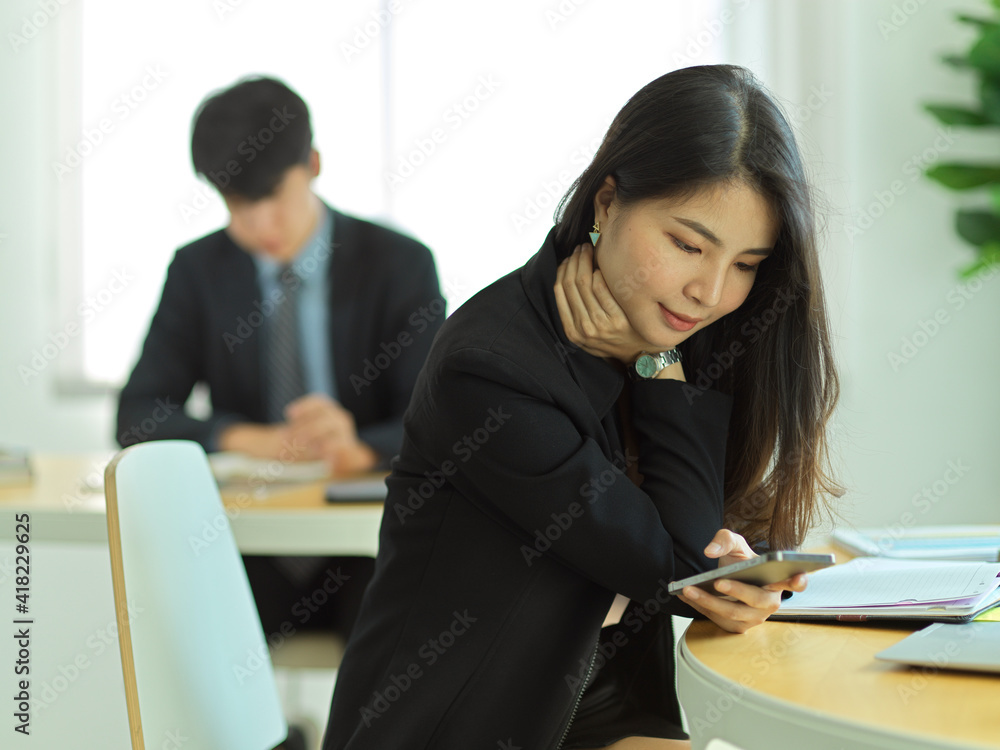 Businesswoman relaxing with smartphone while sitting at workplace with colleague in background Businesswoman relaxing with smartphone while sitting at workplace with colleague in background