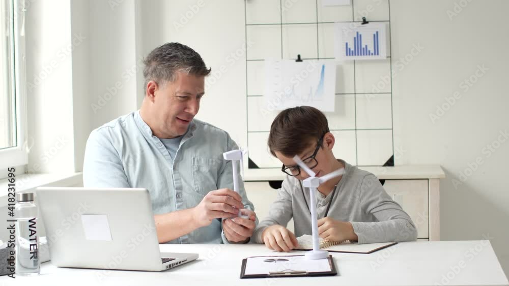 Father and son sitting at table in home office playing and working with two models of wind turbine Father and son sitting at table in home office playing and working with two models of wind turbine