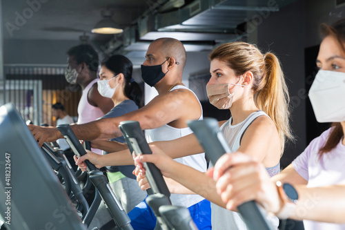 Active people in protective masks having running elliptical trainer class in health club © JackF