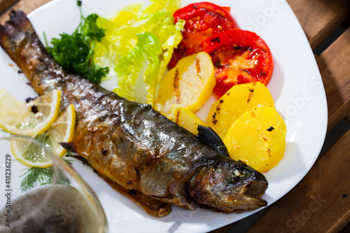 Deliciously baked whole trout with potatoes, tomatoes and greens on white plate, glass of wine