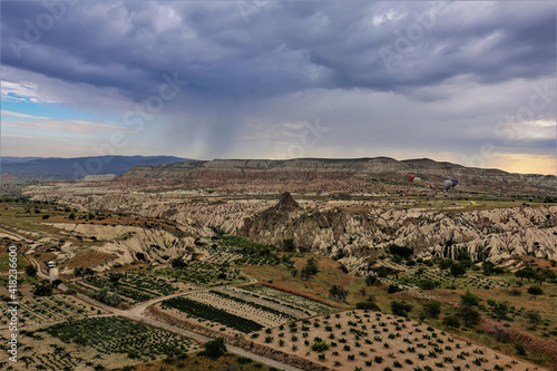 Bird s-eye view of Cappadocia. There are cultivated agricultural fields in the valley. Around - rocks with folded slopes  sharp and flat tops. There is a cloud in the sky  balloons are hovering.