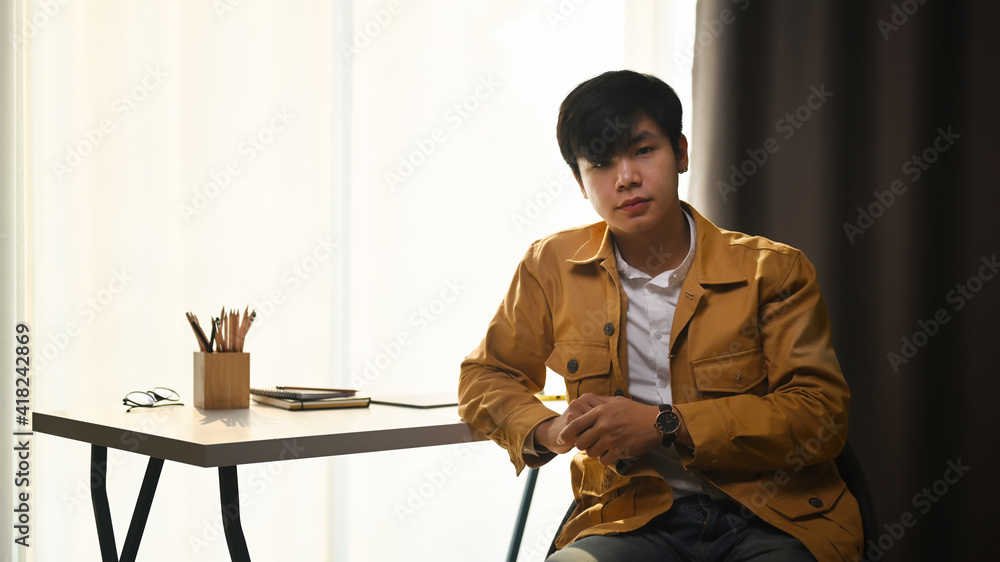Young asian man in yellow jacket looking at camera and sitting near window at his home office.
