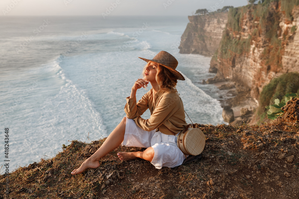 Young woman in straw hat  sitting on cliff and looking at the ocean waves, enjoy vacation on Bali island. Travel Indonesia concept. Happy young woman enjoy summerr holiday